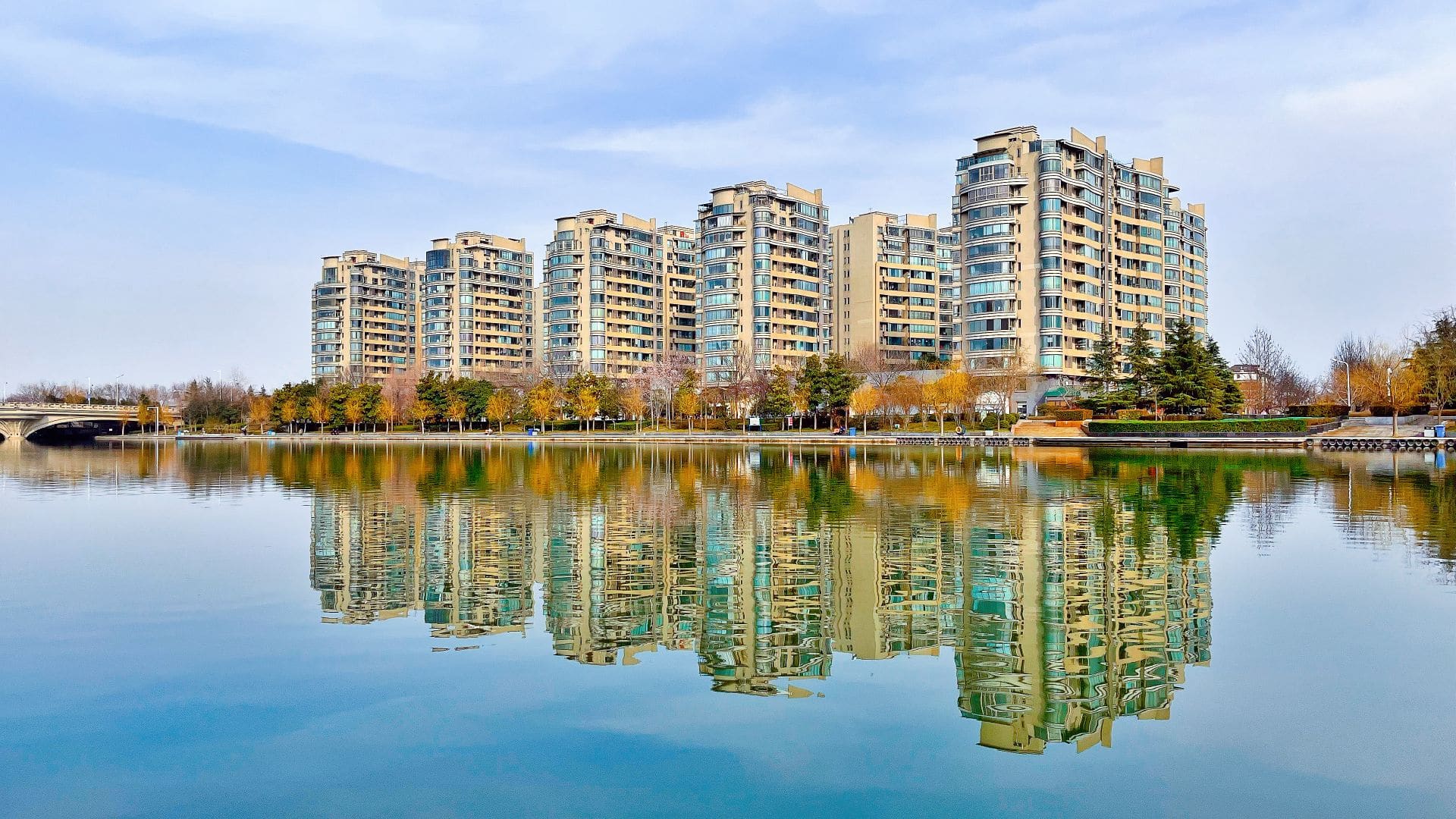 High-rise apartments reflect on a tranquil waterfront in Zhengzhou, China, showcasing urban architecture.
