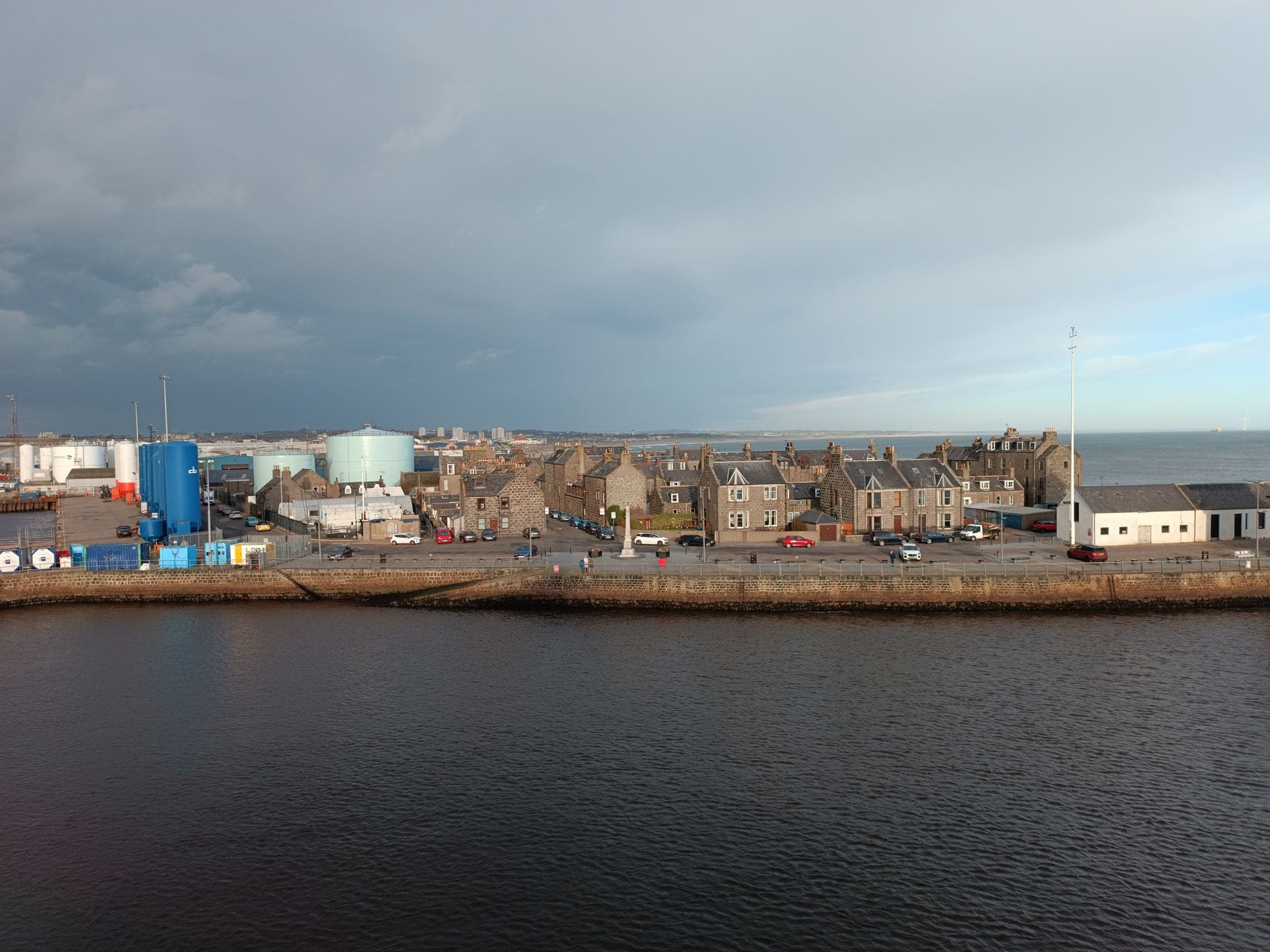 Overview of a coastal town's waterfront with buildings and industrial structures under a cloudy sky.