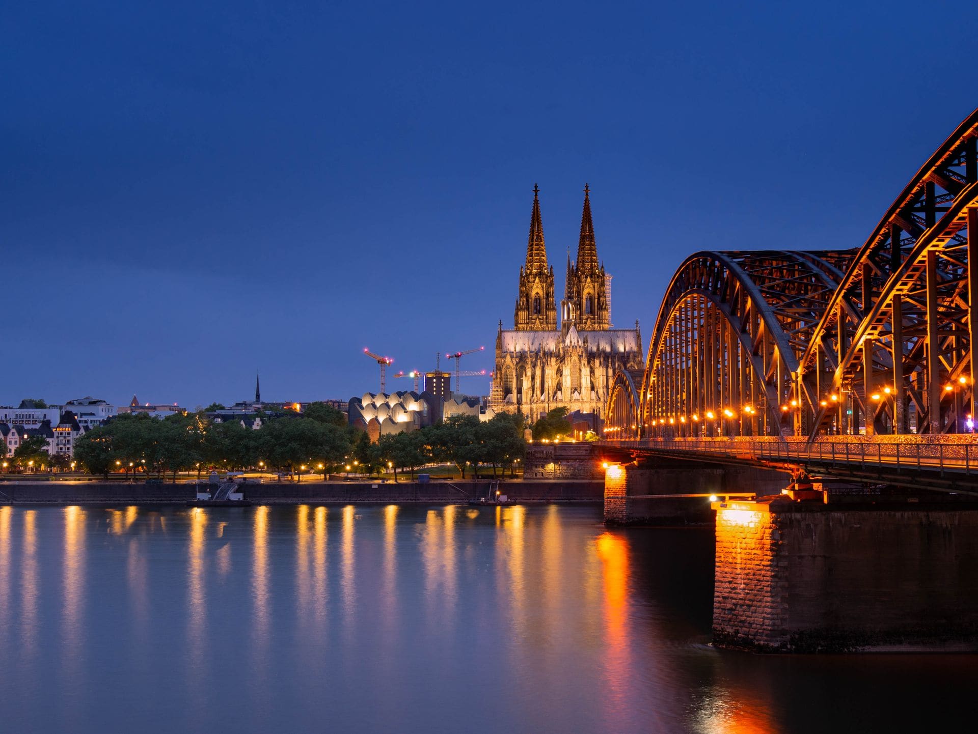 The iconic Cologne Cathedral and Hohenzollern Bridge beautifully lit up against the night sky, reflecting in the Rhine River.