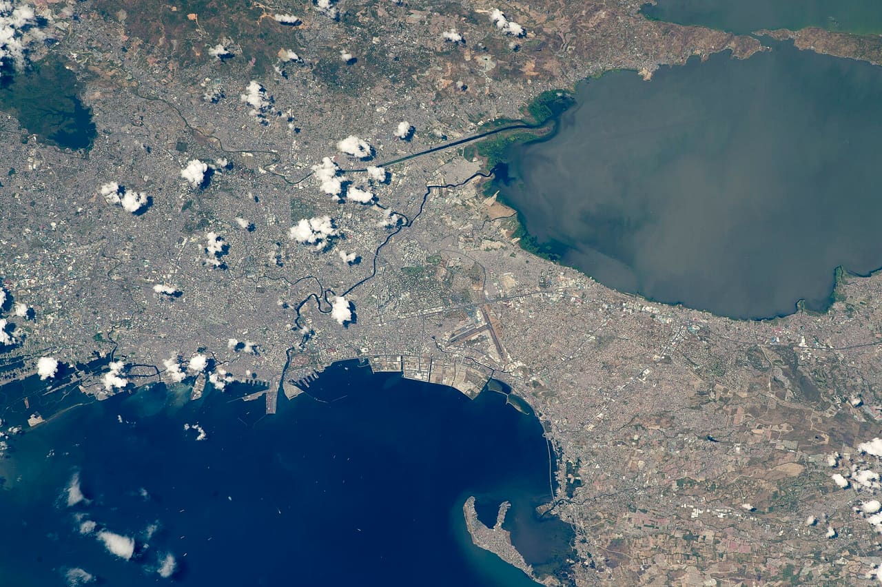 While orbiting over the South China Sea, an astronaut aboard the International Space Station (ISS) took this oblique photograph of the blocky red and gray urban pattern of Manila and surrounding cities. Manila is sandwiched between the salt water of Manila Bay—with its scatter of anchored ships—and Laguna de Bay. The great docks at the port can be seen jutting into Manila Bay (image center left), while the heart of the city lies immediately inland of the port. A forested nature reserve with a lake is located within one of the mountainous areas surrounding the metro zone (top left). The combined population of Manila and neighboring Quezon City is 4.7 million people. Manila itself has one of the highest population densities in the world, with 42,857 people per square kilometer. The metropolitan area is the political and economic hub of the Philippines, and its international trade connections are so widespread the Globalization and World Cities Research Network classed Manila as a global city. Manila’s history has always been connected to trade. Arriving from Mexico, the conquistadors founded Manila in 1571, and soon the city became the Far East anchor of the Acapulco-Manila Galleon trade route connecting Spanish America with Asia. This represents one of the earliest examples of globalization. The dark lines of several waterways are prominent amidst the gray cityscape. The Pasig River winds through the middle of the city. The straight line of the Manggahan Floodway is a flood-control channel engineered to divert water from the Marikina River away from the city center and into Laguna de Bay. Engineering works along the floodway also control tidal saltwater flow into Laguna de Bay. Astronaut photograph ISS047-E-99713 was acquired on May 6, 2016, with a Nikon D4 digital camera using a 400 millimeter lens, and is provided by the ISS Crew Earth Observations Facility and the Earth Science and Remote Sensing Unit, Johnson Space Center. The image was taken by a member of the Expedition 47 crew. The image has been cropped and enhanced to improve contrast, and lens artifacts have been removed. The International Space Station Program supports the laboratory as part of the ISS National Lab to help astronauts take pictures of Earth that will be of the greatest value to scientists and the public, and to make those images freely available on the Internet. Additional images taken by astronauts and cosmonauts can be viewed at the NASA/JSC Gateway to Astronaut Photography of Earth. Caption by M. Justin Wilkinson, Texas State University, Jacobs Contract at NASA-JSC.