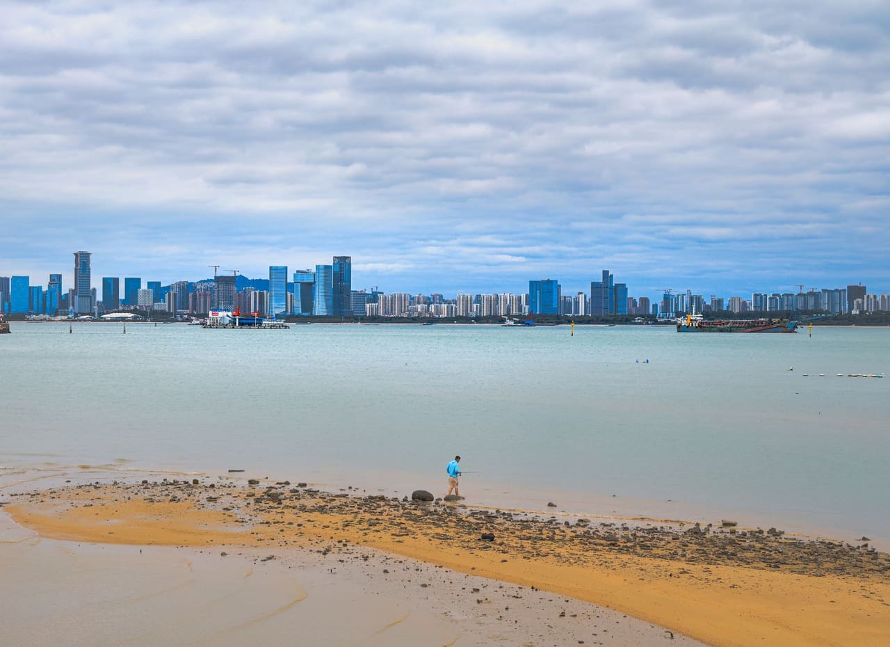 A distant skyline of Haikou, China viewed from a beach, with one person walking on the sand.