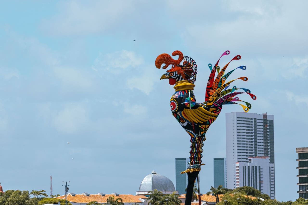 Vibrant rooster sculpture overlooking Recife skyline, capturing Brazilian culture.