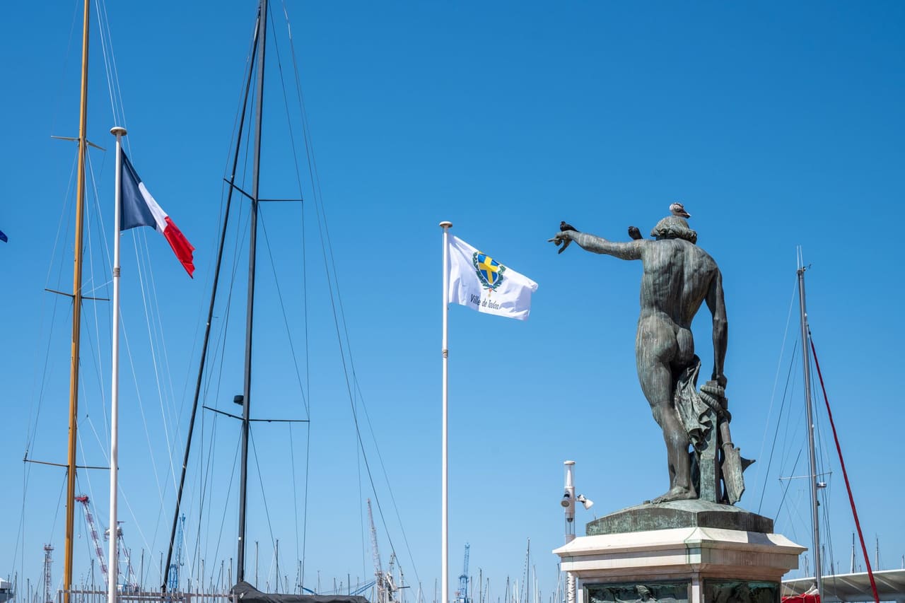 A statue overlooks sailboat masts with flags in Toulon Harbor, France under a clear blue sky.